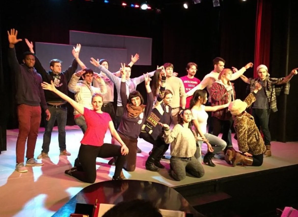 Group of diverse performers on stage during a theater or dance rehearsal, some kneeling and others standing, with stage lighting and a dark background.