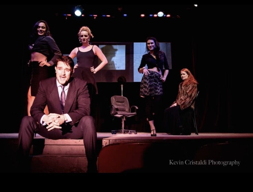 Five actors on stage in a theatrical play, with one man in the foreground sitting on the edge of the stage and four women standing behind him, dressed in black outfits, with a black chair and a monitor in the background, under stage lighting.