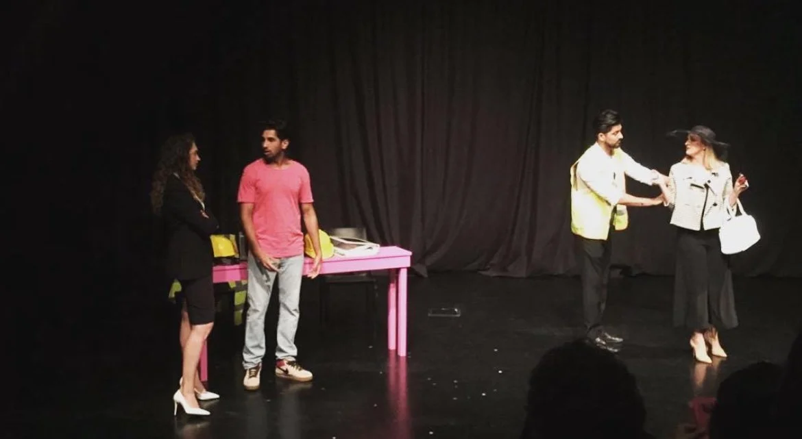 Four people on stage with black curtains in the background; two are near a pink table, and two are engaging in a handshake or conversation.
