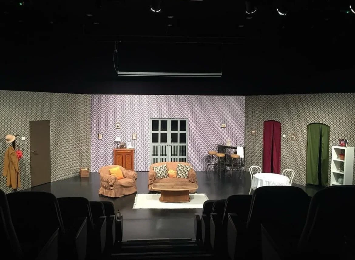 Theater stage setup with vintage furniture including a sofa and armchair, patterned wallpaper, small tables, and colorful room dividers, ready for a performance.