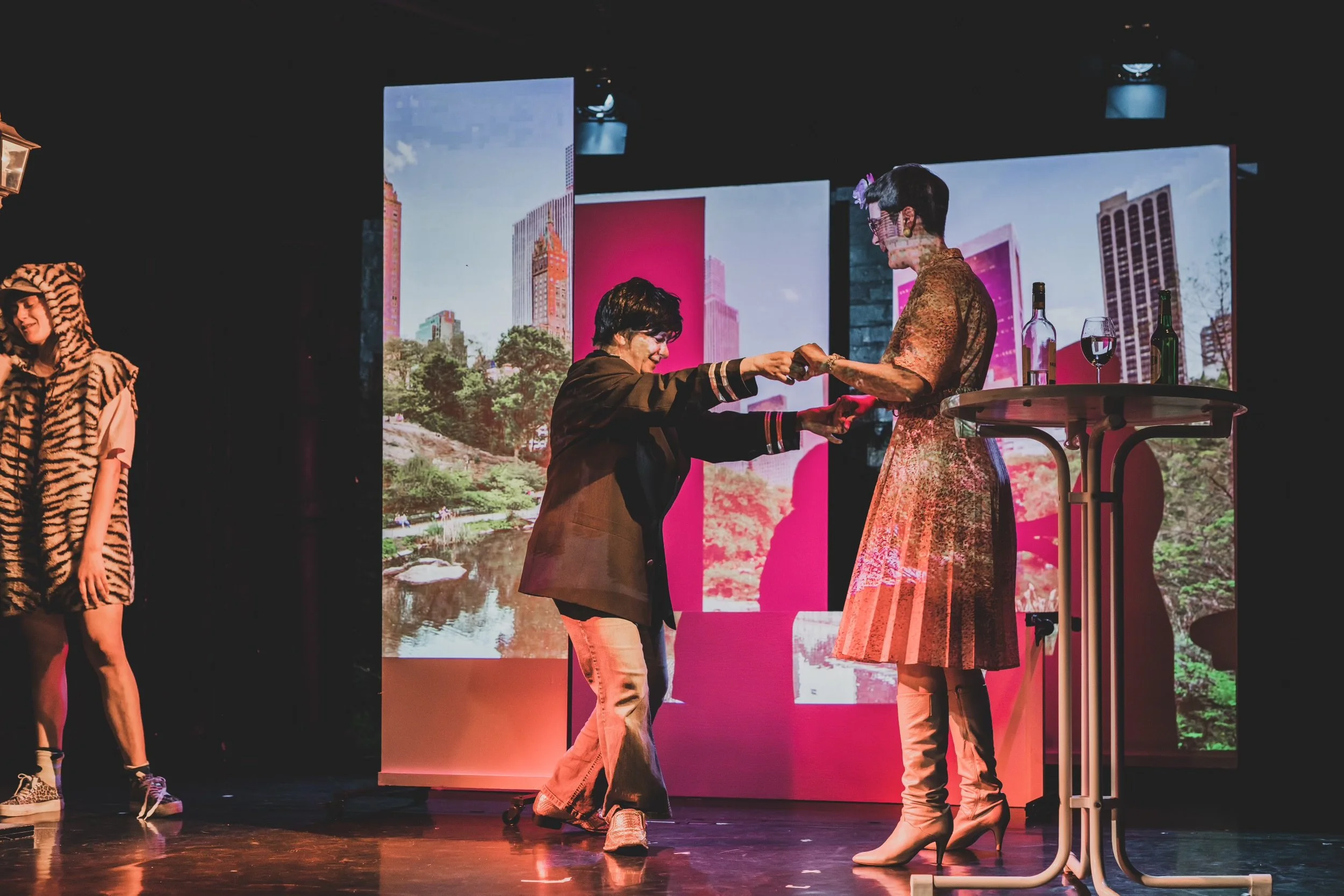 Two women on stage, one handing a glass to the other, with a backdrop of cityscape images on screens. One woman is dressed in a colorful, patterned dress with high heels, and the other is in a dark jacket and beige pants. A third person in a zebra costume is partially visible on the left side.