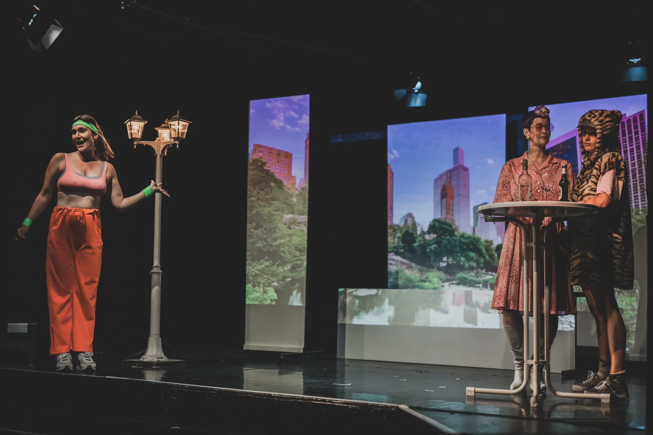 Three women on stage, two standing at a table with bottles, one on the left in exercise attire near a lamppost, with cityscape screens in the background.