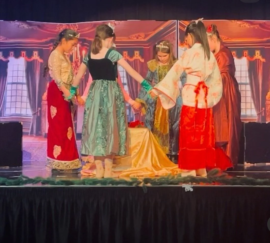 Children dressed in colorful costumes performing a play on stage with a royal or historical setting background.