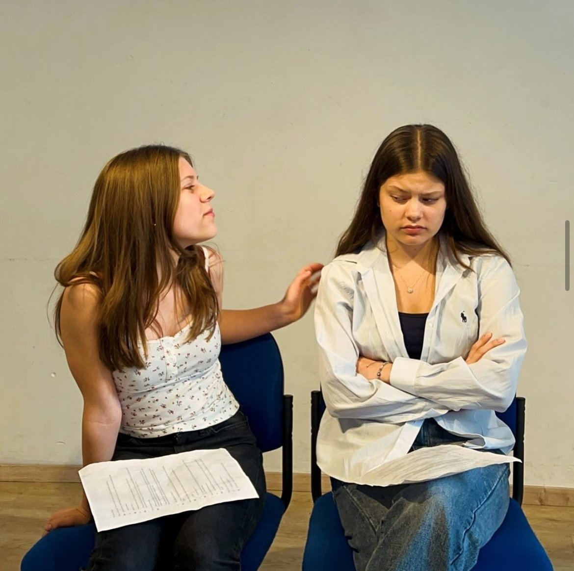 Two young women sitting on chairs, one comforting the other with arms on her shoulder, in a room with plain light-colored wall.