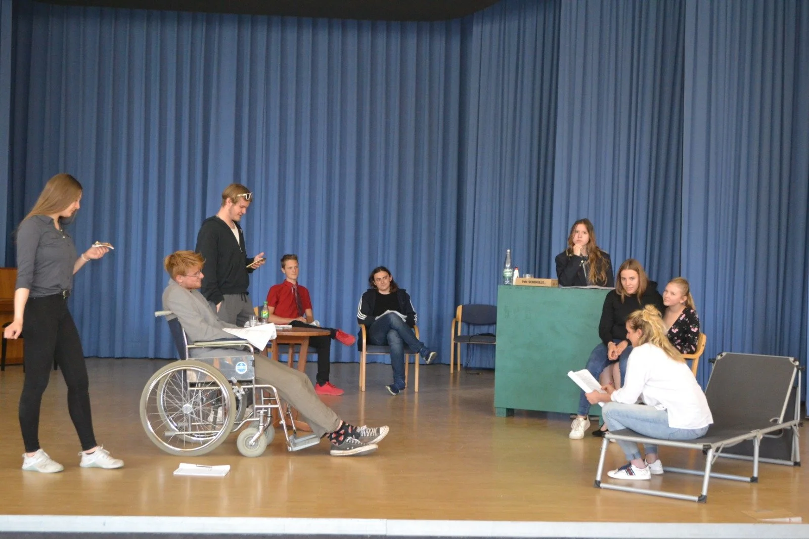 Group of people participating in a staged play or scene in front of blue curtains, including a person in a wheelchair, seated at a desk, and others sitting or standing, engaged in acting or reading roles.