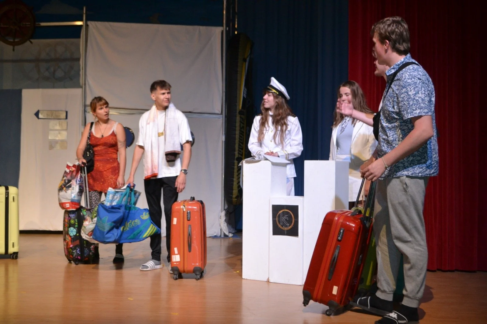 A group of young people standing on a stage with suitcases, waiting to check in at a travel counter, with three people behind the counter dressed as airline or travel staff.