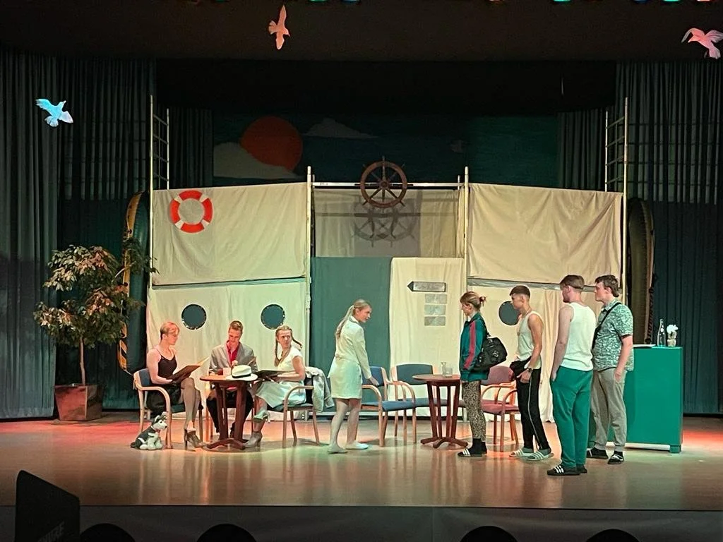 Theater scene with actors on stage portraying a cruise ship setting, with decorations like a ship's wheel, lifebuoy, and seagull cutouts.