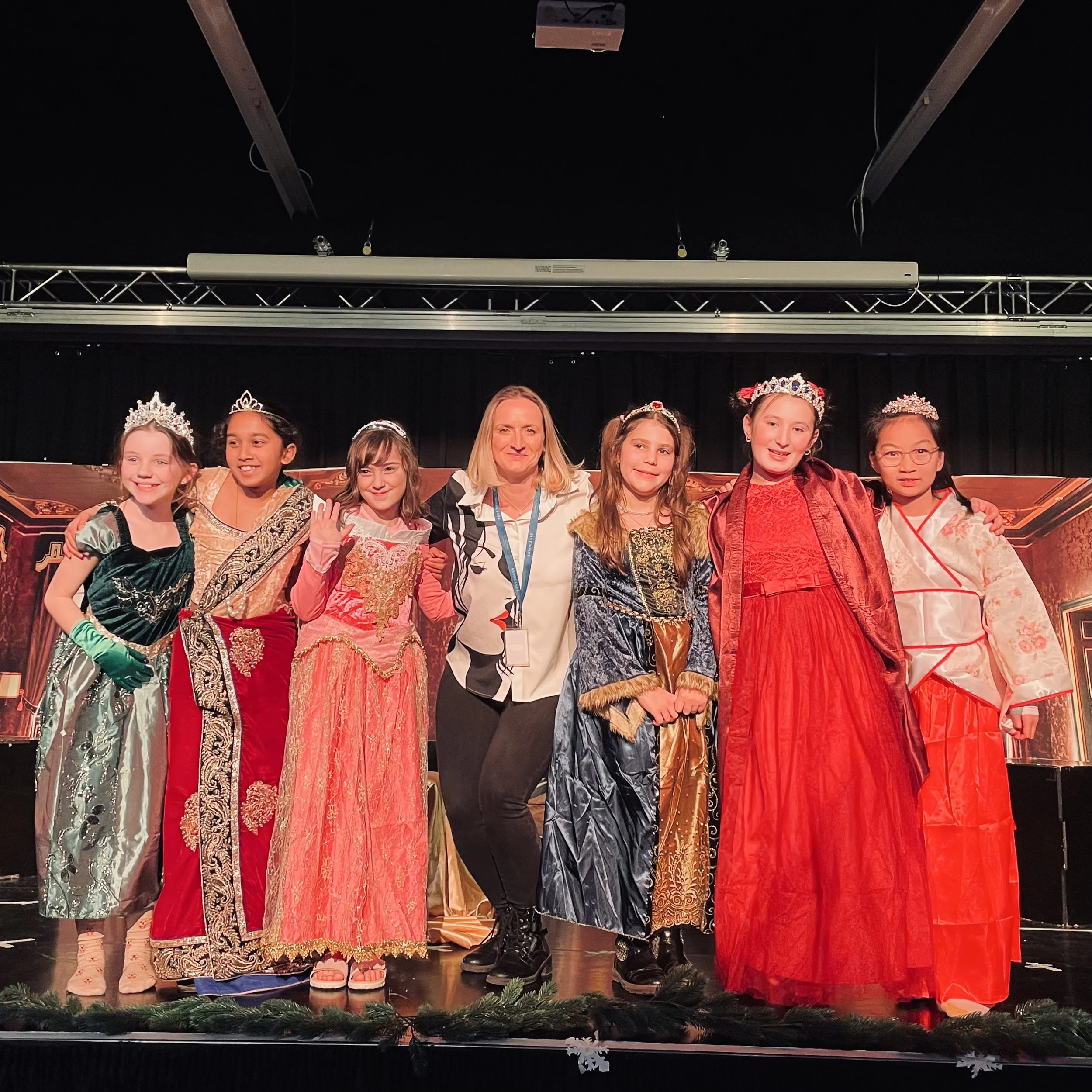 Seven young girls dressed in princess costumes with a woman in the center, on a stage with a backdrop of an ornate room, wearing tiaras and crowns, during a costume event or performance.