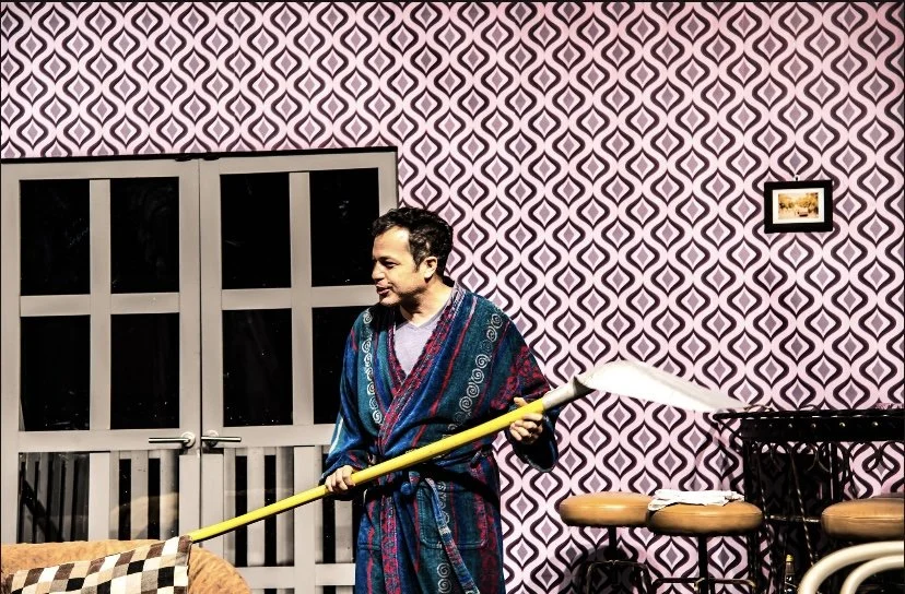 A man standing indoors, wearing a colorful bathrobe, holding a mop, with patterned wallpaper and a closed door in the background.