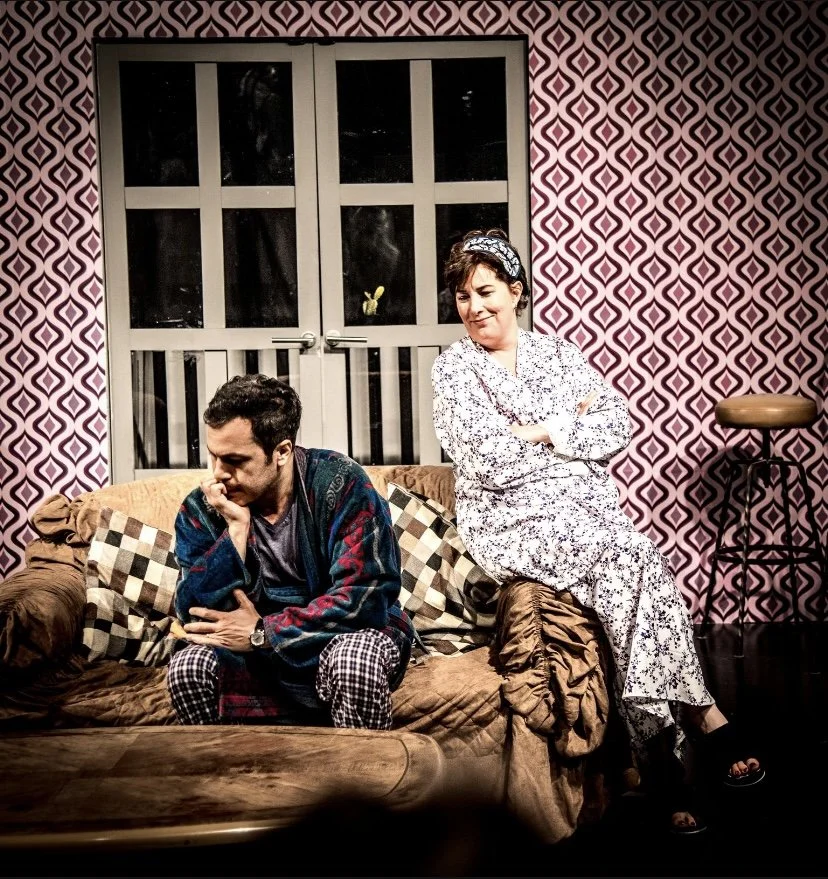 Two people sitting on a sofa in a room with pink and black patterned wallpaper, dressed in pajamas, with one person looking down thoughtfully and the other person smiling with arms crossed.