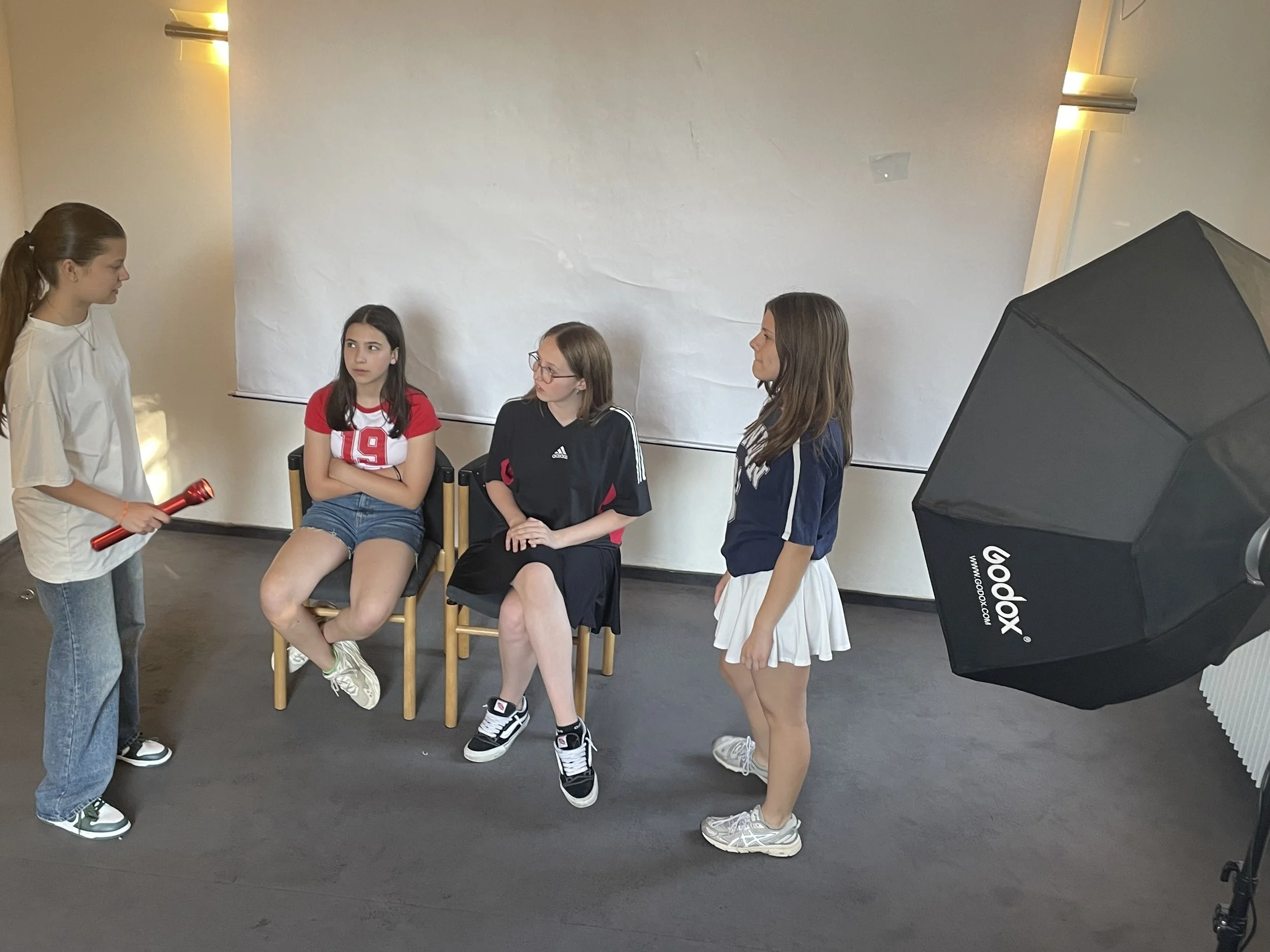 Four girls in a photo shoot, with one girl standing on the left holding a red light, and three girls sitting or standing, with two seated on chairs and one standing, against a plain white backdrop, with a large studio light on the right side.
