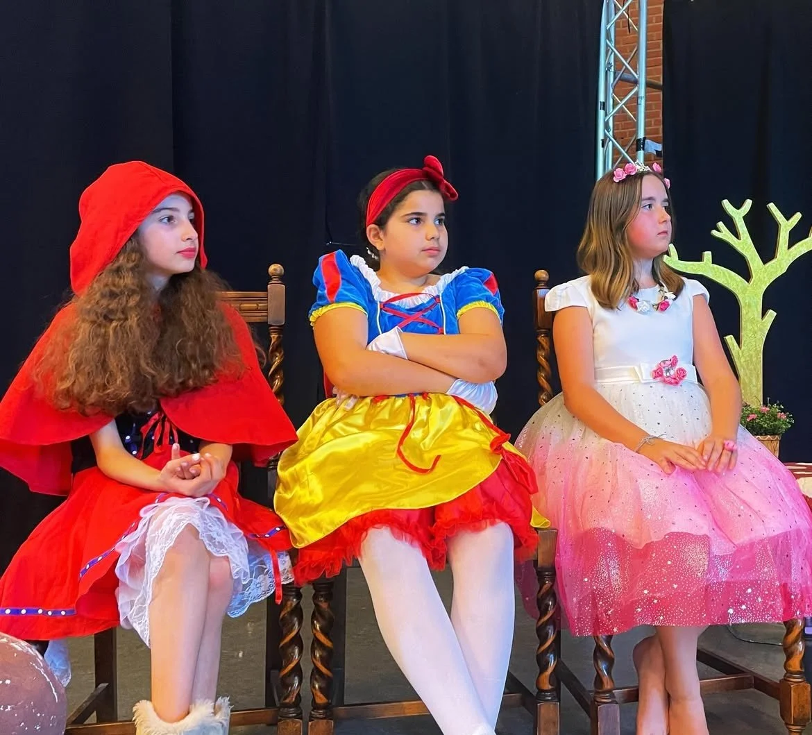 Three young girls dressed in fairy tale costumes sitting on chairs during a performance, with dark background and decorative props.
