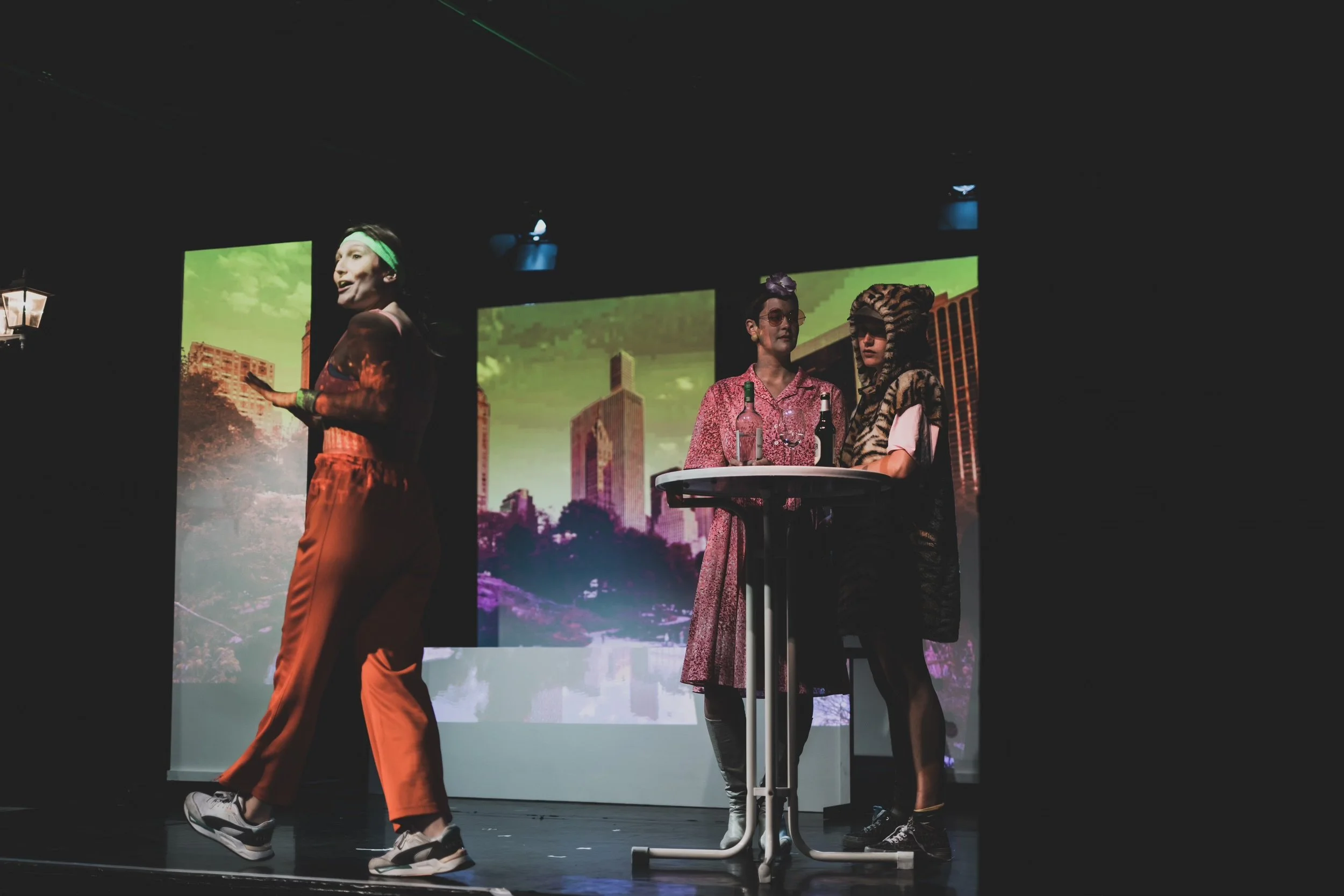 Three women in costumes performing on stage with cityscape images projected behind them. One woman in bright orange pants and a green headband is in motion, and two women are near a table holding bottles, one dressed in a vintage pink dress and the other in animal print with a hood.