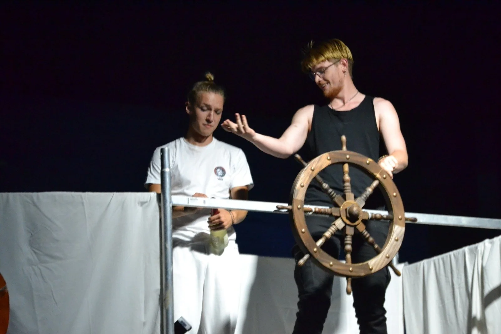 Two young men on a stage, one with long blond hair and a black tank top, and the other with short blond hair and a white shirt, working with a ship's wheel prop.