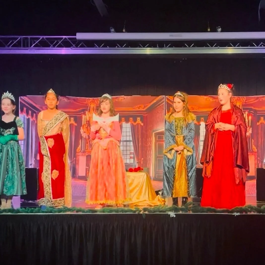 Children dressed in regal costumes, performing a play on a stage with a backdrop of an ornate royal interior.