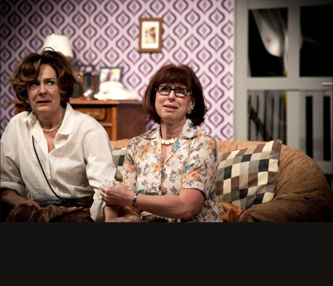 Two women sit on a couch, one looks surprised while the other appears emotional, in a room with a patterned purple wallpaper, a lamp, picture frames, and a sliding glass door in the background.
