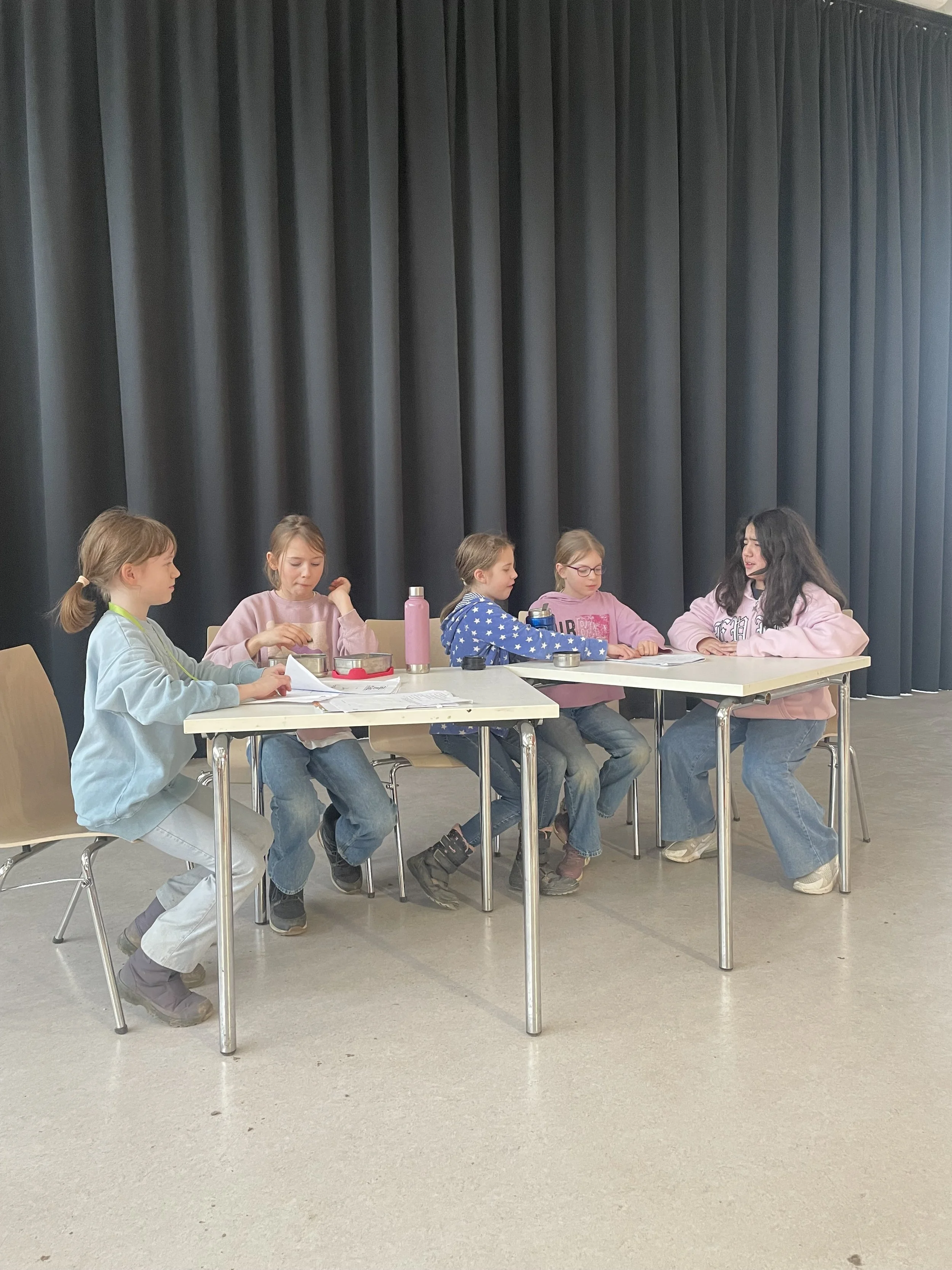 Five young girls sitting at tables on a stage behind a black curtain, engaged in conversation or activities.