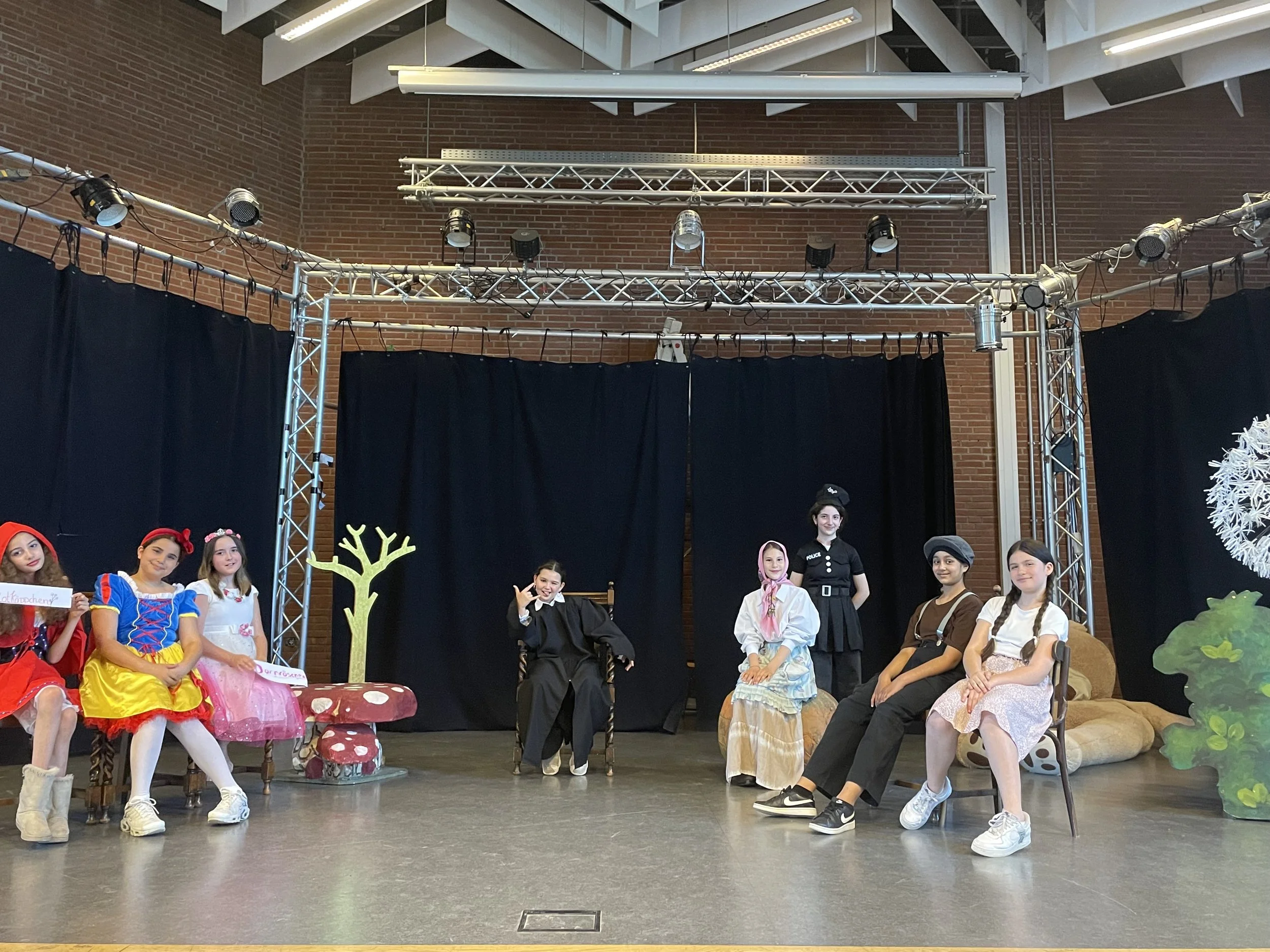 Children performing a play on a stage with black curtains, decorated with props like a tree, a mushroom, and greenery, some children are dressed in costumes like Snow White, and others are in different costumes, with a black backdrop and stage lighting above.