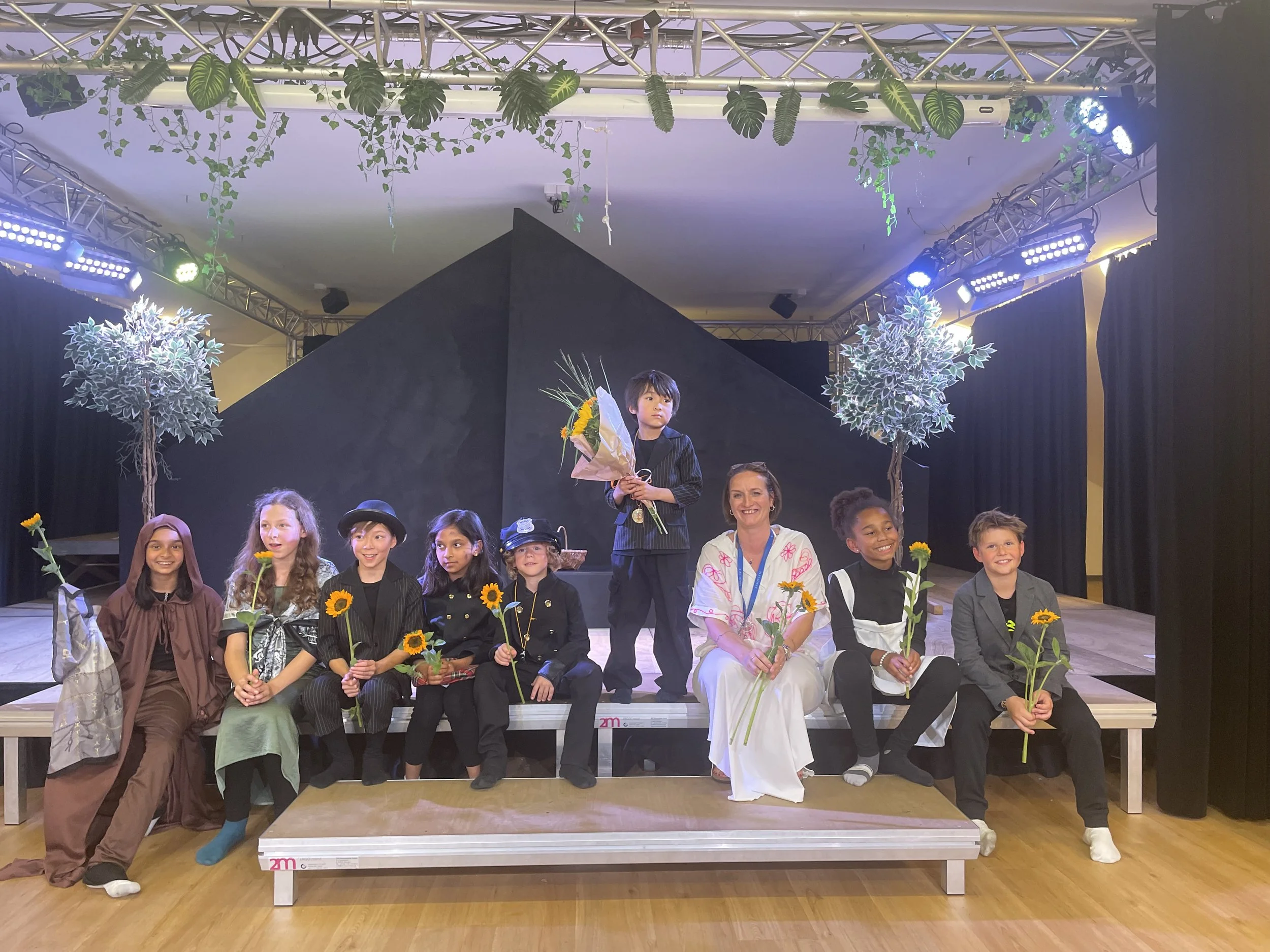 Children and a woman sitting and standing on a stage holding yellow flowers, with a black geometric backdrop, stage lights, and hanging plants overhead, celebrating a performance or event.
