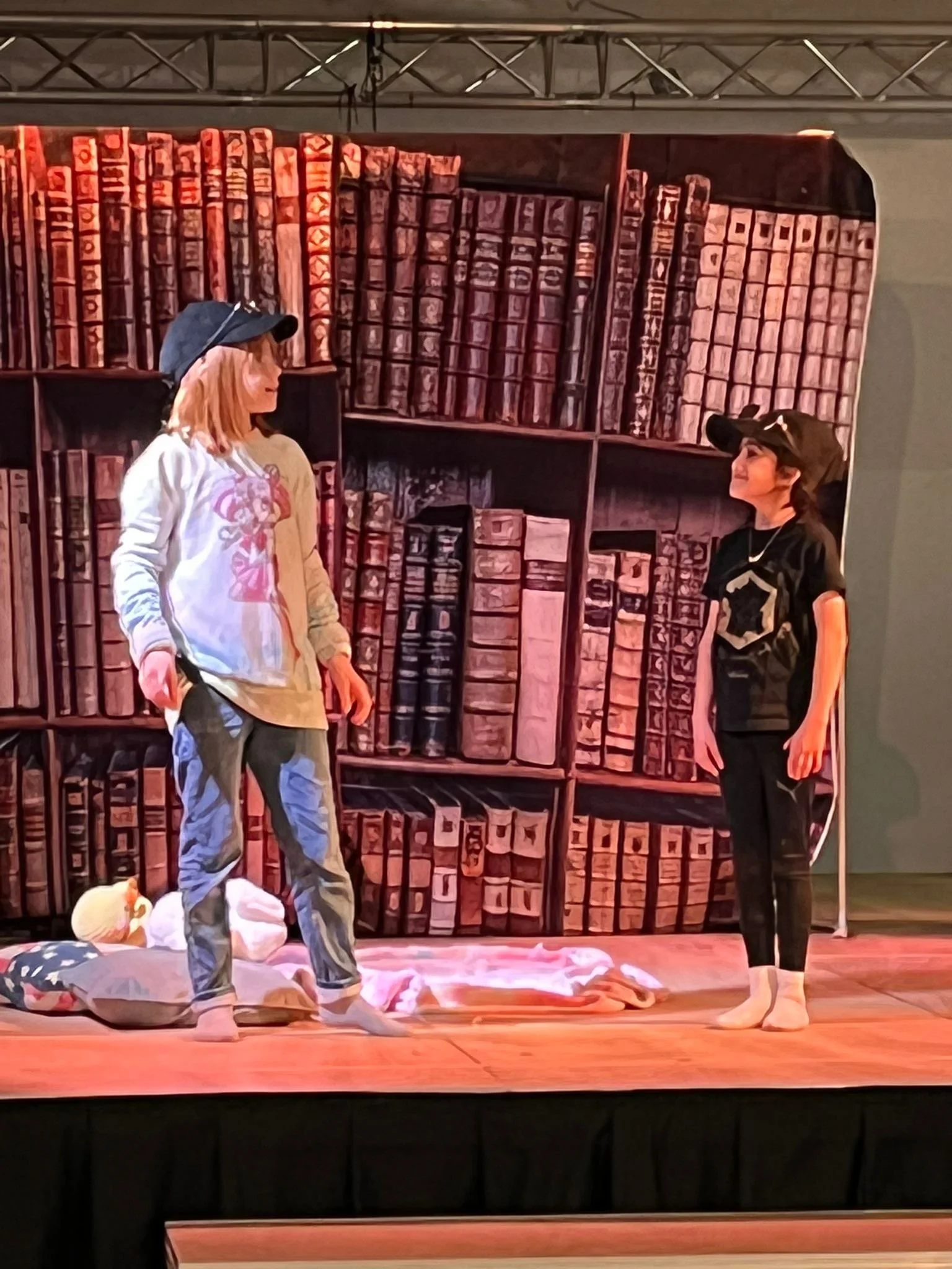 Two women standing on a stage with a bookshelf backdrop, one wearing a gray hoodie and the other in black, engaging in a conversation. There are belongings on the floor, including a pillow and a blanket.