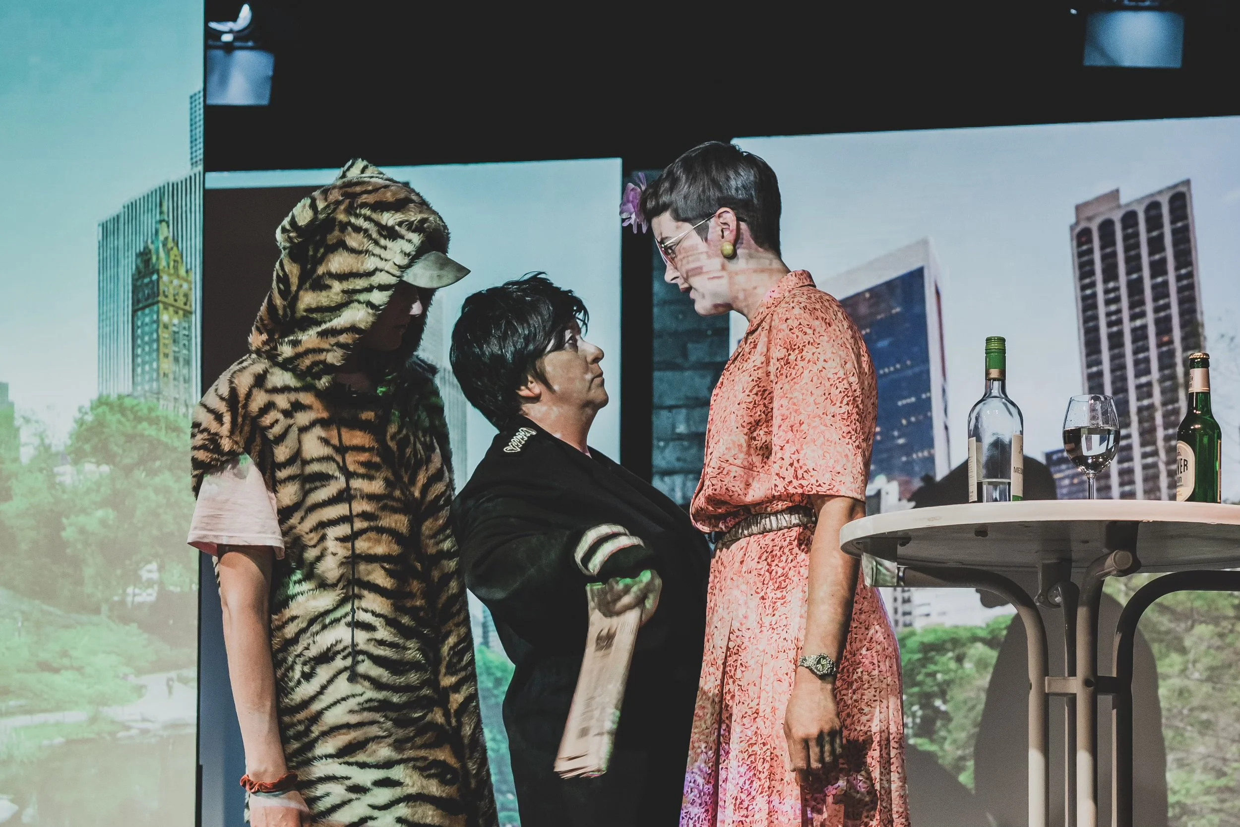 Three women standing close together, two facing each other and one turned to the side, with a cityscape background on a large screen.