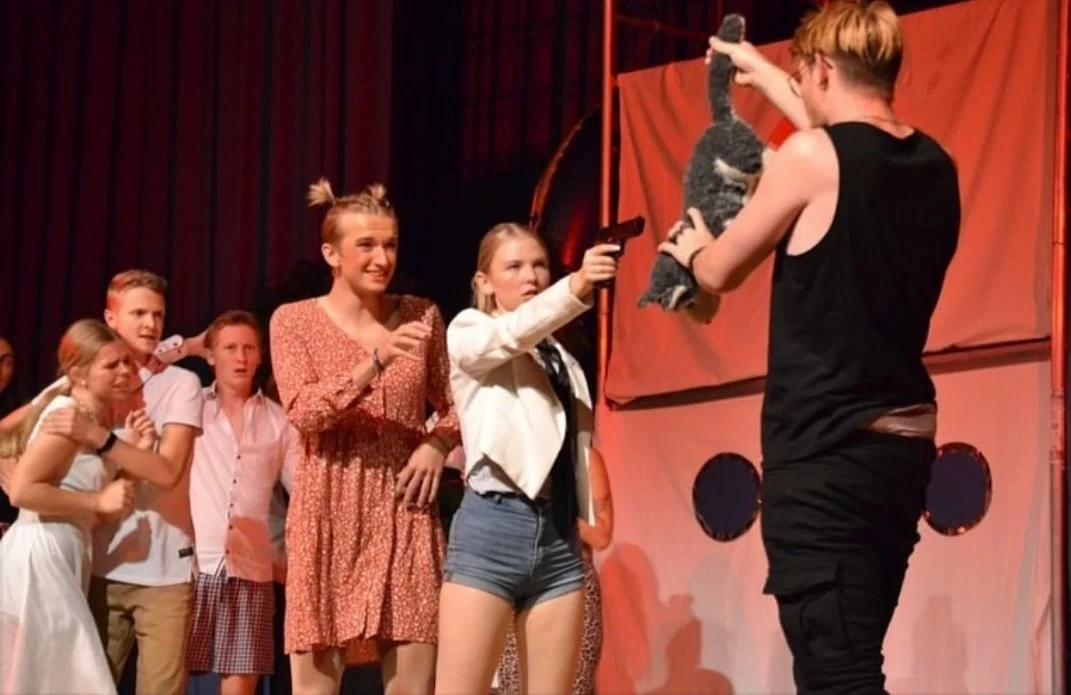 A group of children on stage at a puppet show, with one girl holding a gun prop and pointing it at a performer holding a cat puppet, as others watch in surprise or excitement.