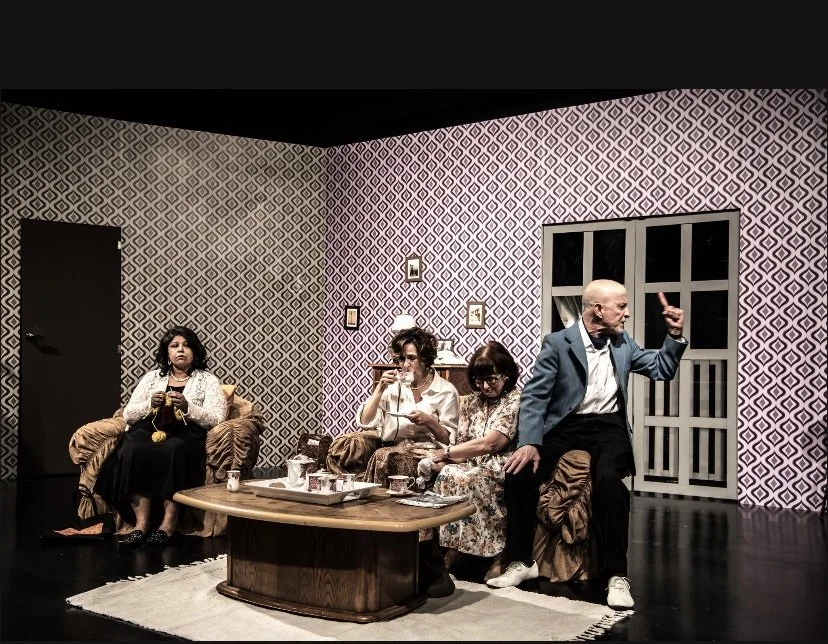 Four people sitting in a living room with patterned wallpaper, one man standing and talking, three women sitting, one drinking from a cup. The room has a window, a coffee table with cups, and a rug.