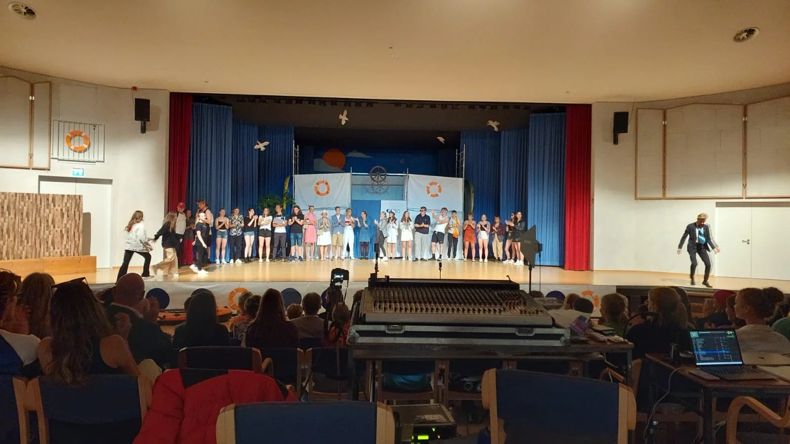 A group of performers on stage during a theatrical production, with a painted backdrop of a nautical scene and props resembling parts of a ship, and audience members watching from seats in a theater.