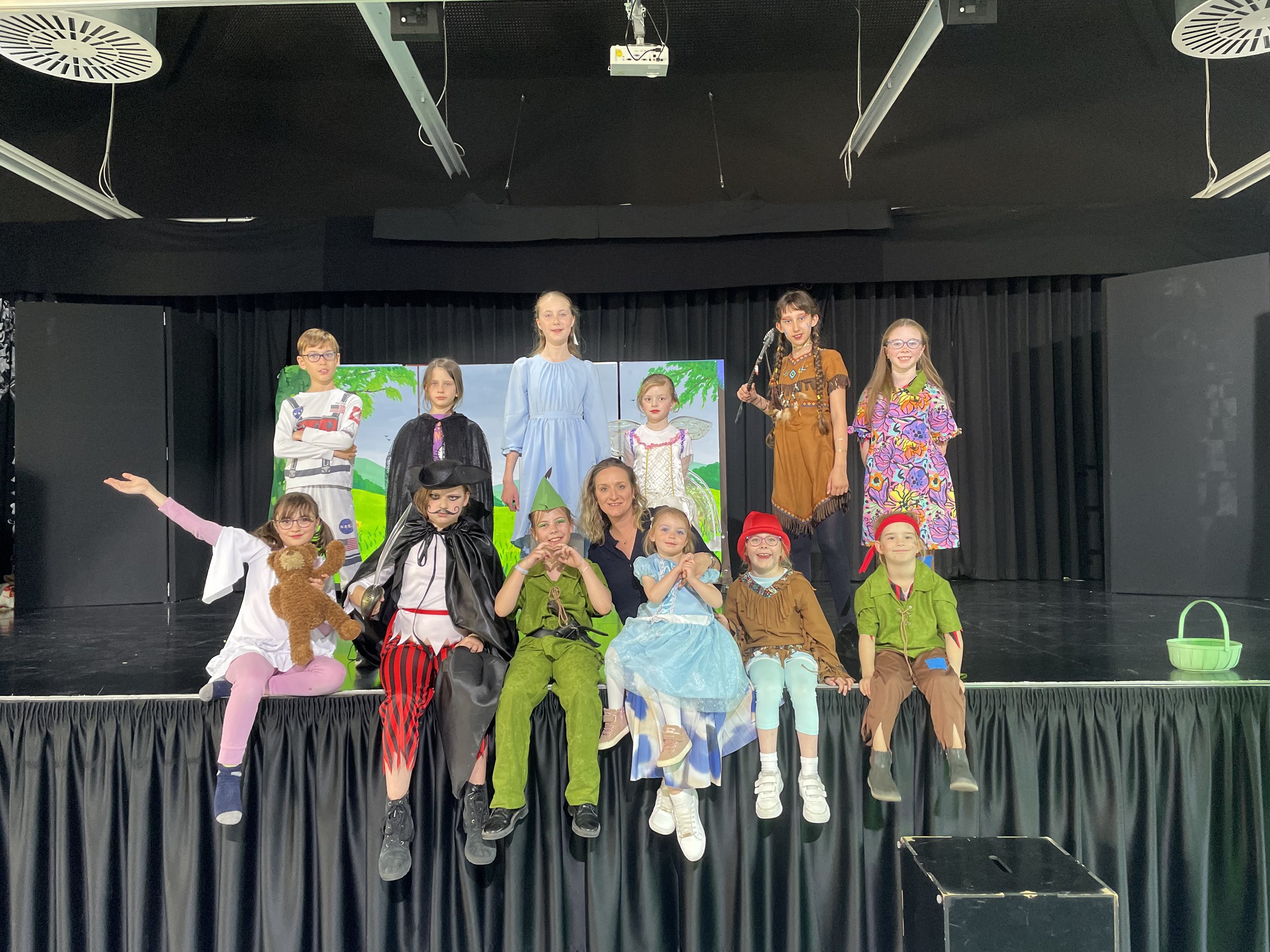 Children dressed in colorful costumes performing on stage, with some sitting and others standing, in front of a backdrop depicting a green landscape with trees.
