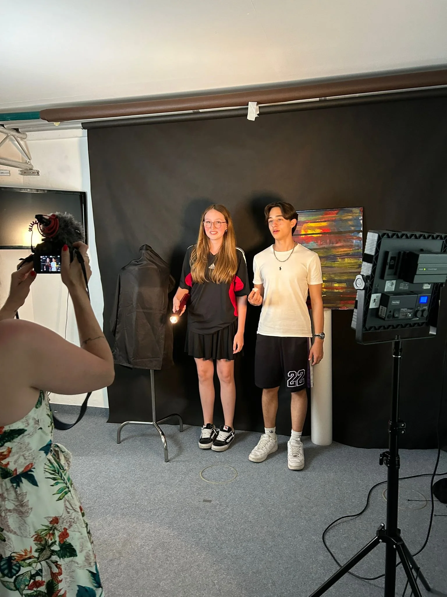 Two young people, a girl with long hair and glasses in a black and red sports jersey and a black skirt, and a boy with dark hair in a white t-shirt and black shorts, stand together in front of a black backdrop, being photographed in a studio with professional lighting. A person in a floral dress is taking their picture with a camera.