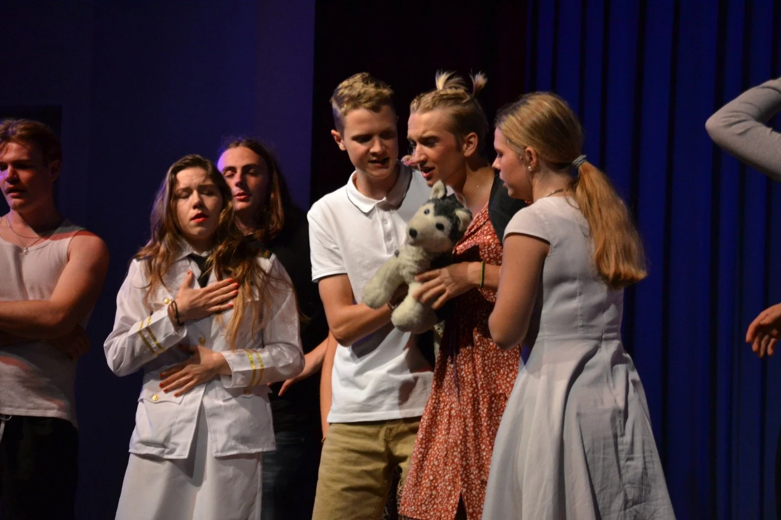 Group of young people on stage performing a theatrical scene, with one holding an animated plush husky dog.