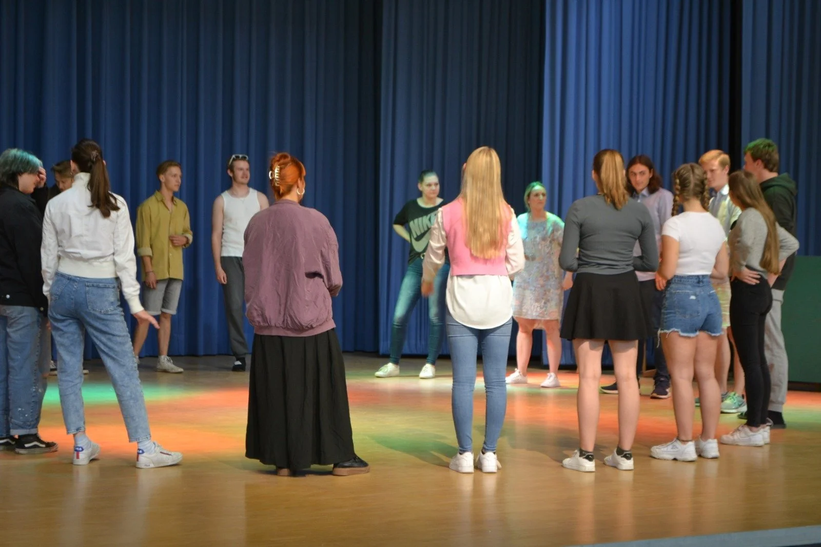 Group of young people gathered on stage in front of blue curtains, possibly rehearsing or performing.