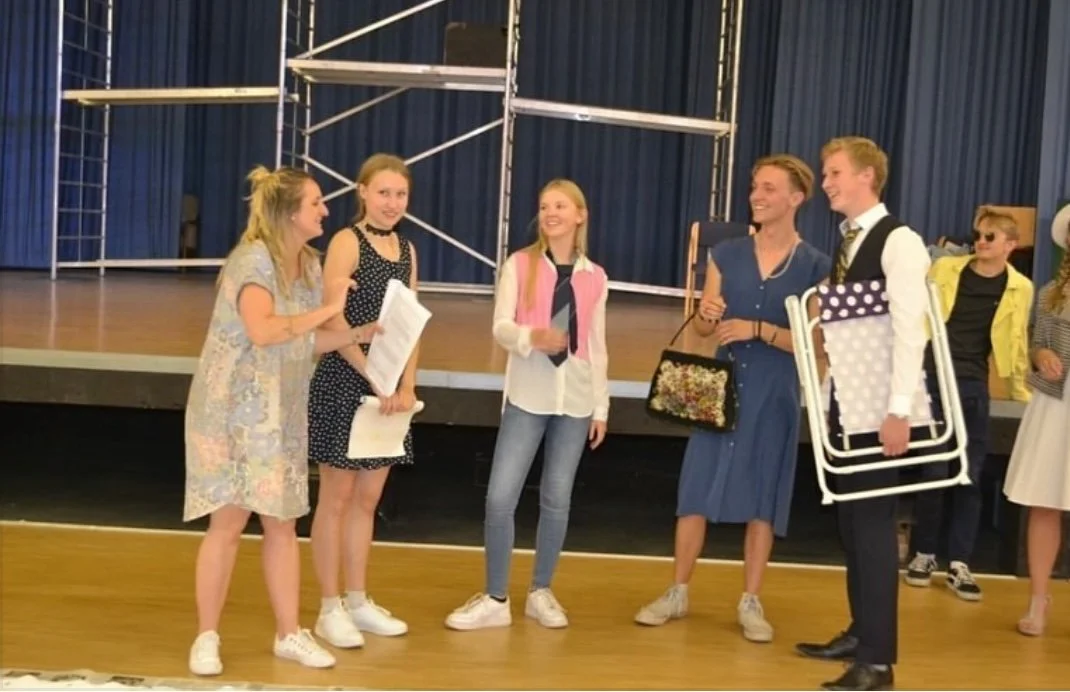 Group of teenagers standing on stage, engaging in conversation, with blue curtains and scaffolding in the background.