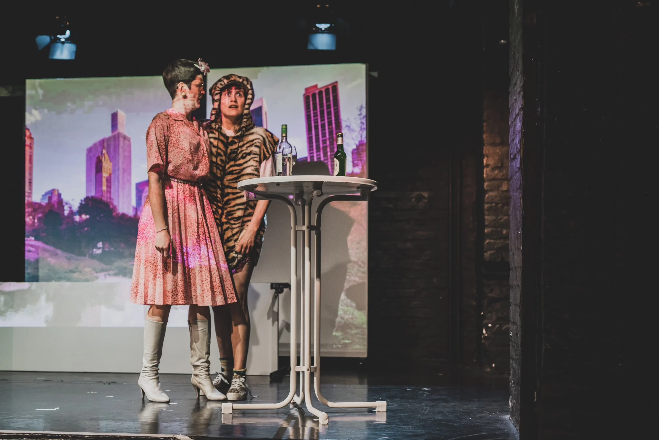 Two women dressed in retro costumes stand on a stage near a high-top table with two bottles on it, performing in front of a cityscape backdrop.