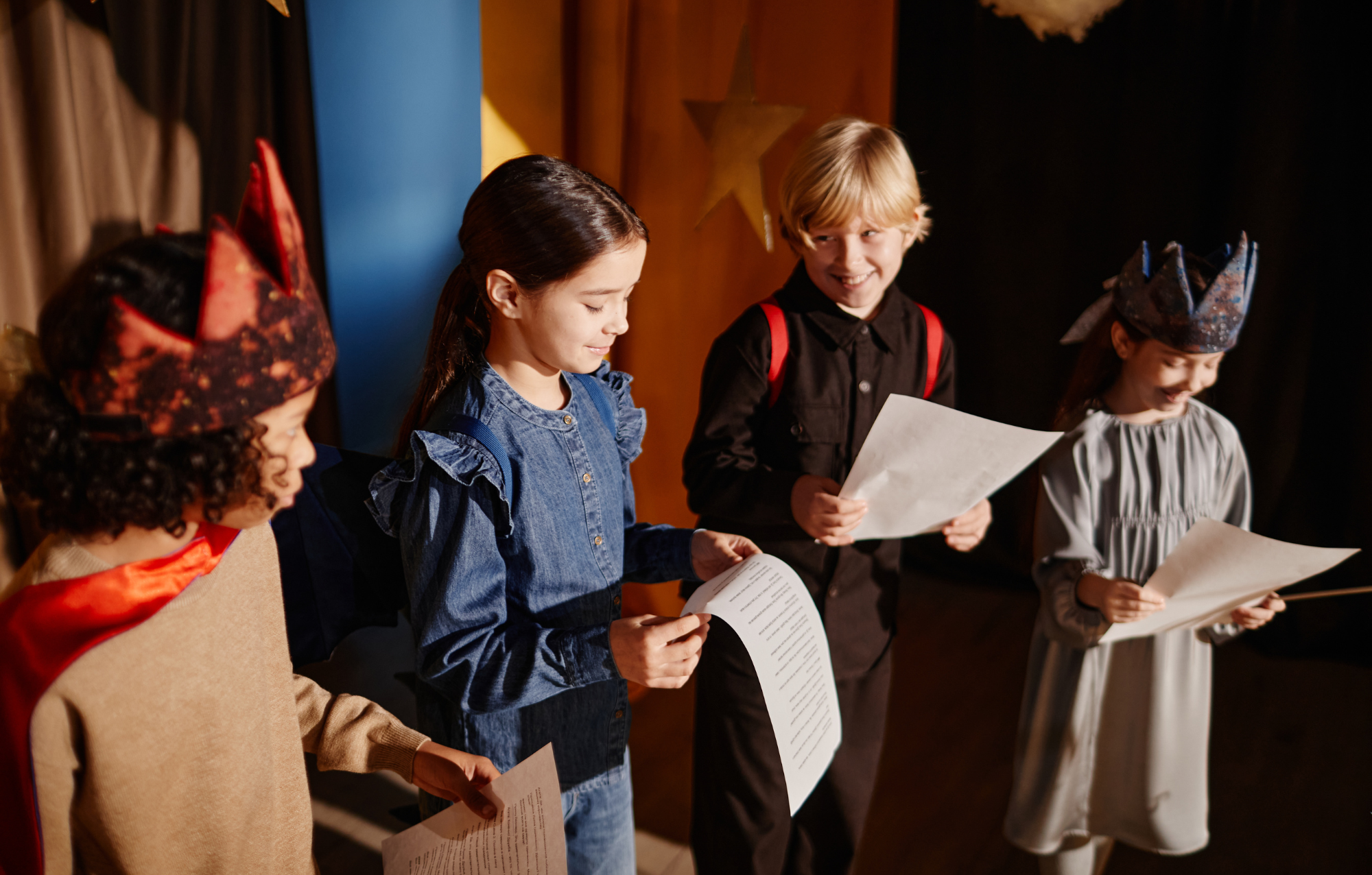 Children on stage reading from scripts, some wearing colorful crowns, during a school performance.