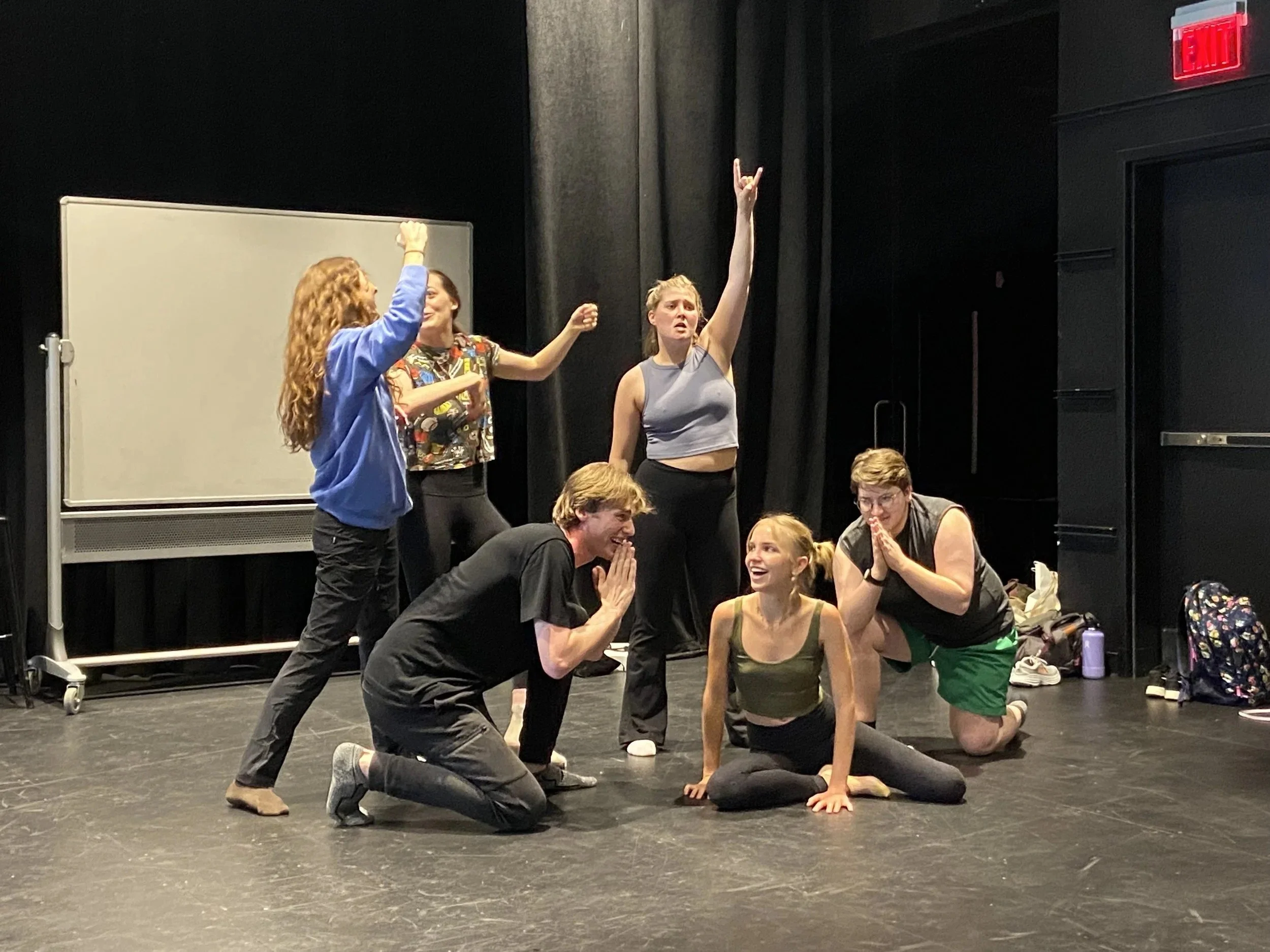 Group of six young people acting or rehearsing on a stage, with various expressive poses, some kneeling or sitting, and a whiteboard and black curtains in the background.