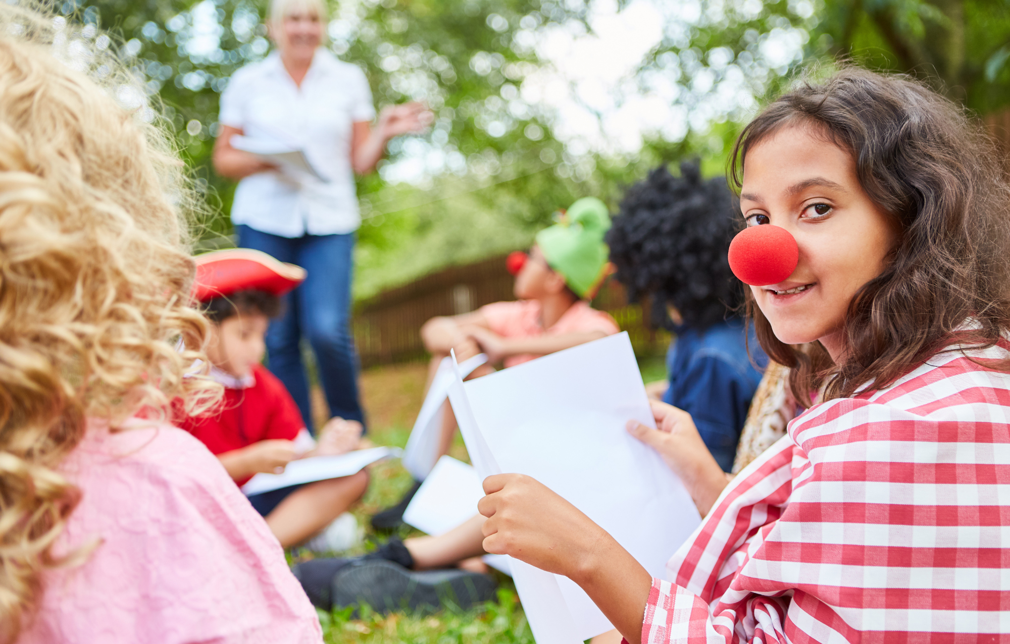 Girl with a red clown nose and a red-and-white checkered shirt holding papers in an outdoor setting with other children dressed in costumes and a woman standing in the background.