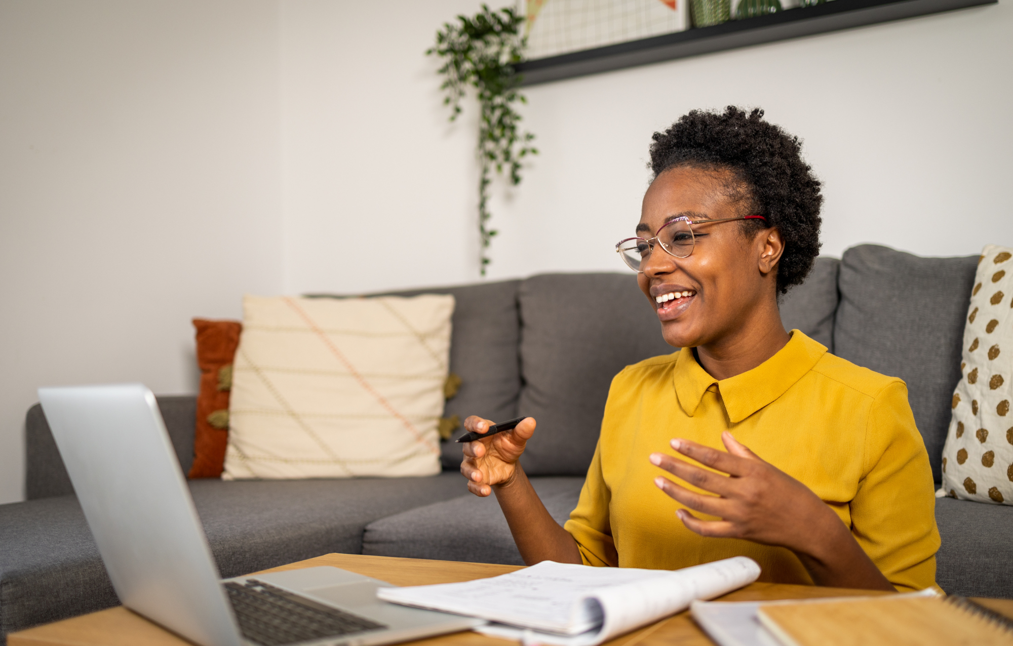A woman with short curly hair and glasses, wearing a yellow blouse, sitting at a table with a laptop and papers, smiling and gesturing with her hand during a video call in her living room.