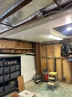 Garage interior with exposed wooden framing, a workbench, and storage shelves, illuminated by overhead lighting.