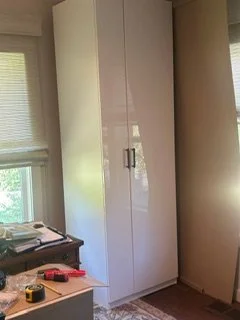 White wardrobe next to a beige column in a room with windows and various items on a desk.