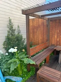 Outdoor wooden pergola with bench seating on a wooden deck, surrounded by green plants and a wooden fence.