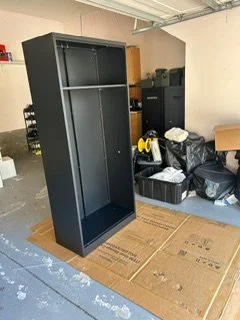 Empty black metal shelving unit in a garage or storage room with cardboard on the floor and various storage items in the background.