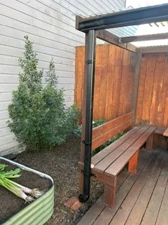 Outdoor wooden patio corner with a brown wooden bench, a wooden privacy fence, and a metal or plastic downspout in the corner. There's a planter with green plants nearby.