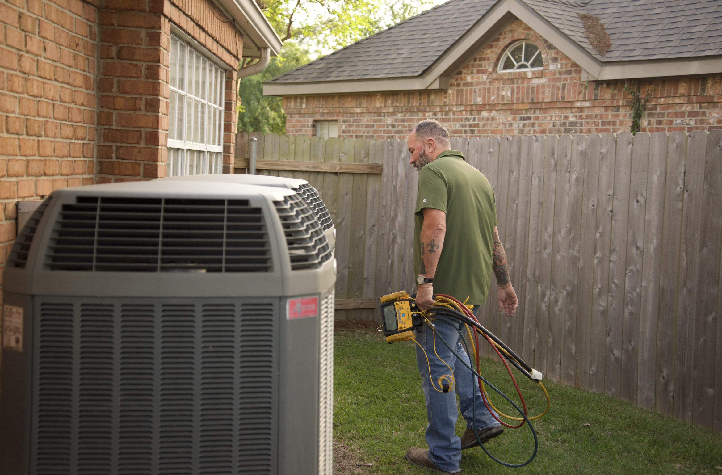 A man working outside next to an air conditioning unit, holding electrical tools and cables, with a wooden fence and brick houses in the background.