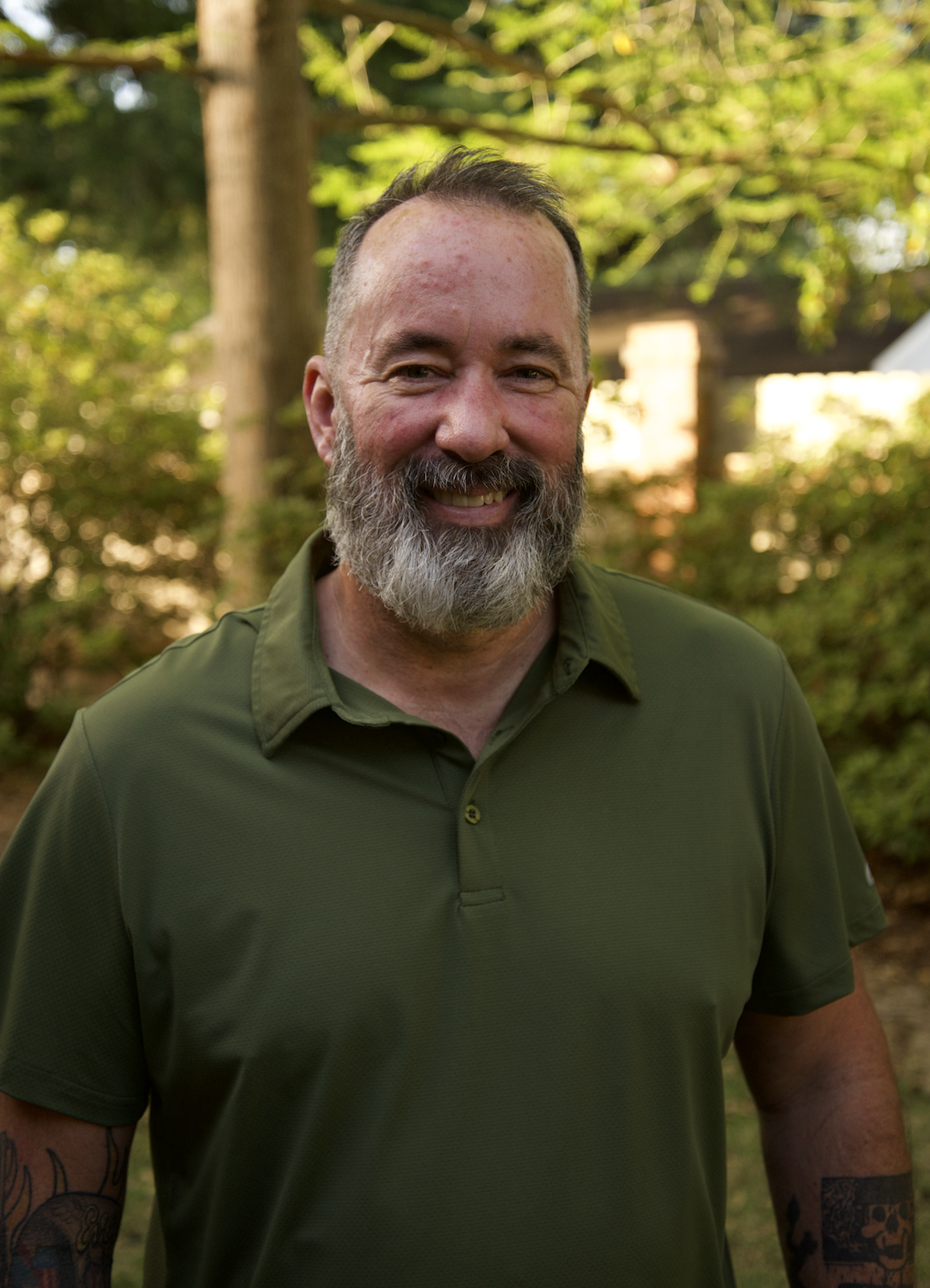 A smiling man with a beard, wearing a green polo shirt, outdoors in a wooded area with trees and sunlight in the background.