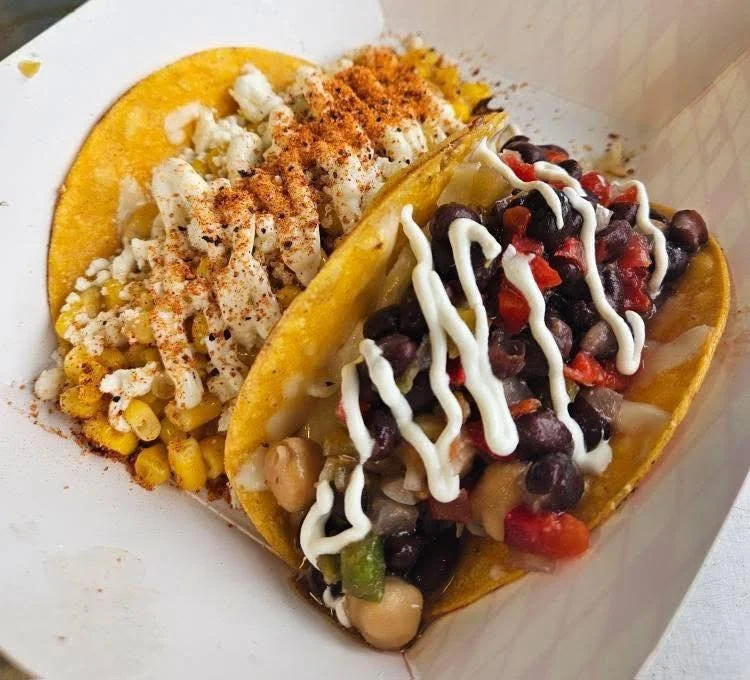 Two hard shell tacos filled with beans, vegetables, and topped with mayonnaise and salsa, served with a side of corn and shredded cheese.