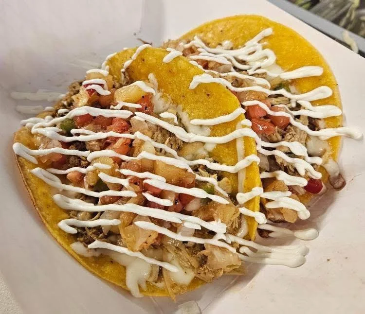 Two tacos with yellow shells filled with meat, vegetables, and topped with sour cream in a white container.