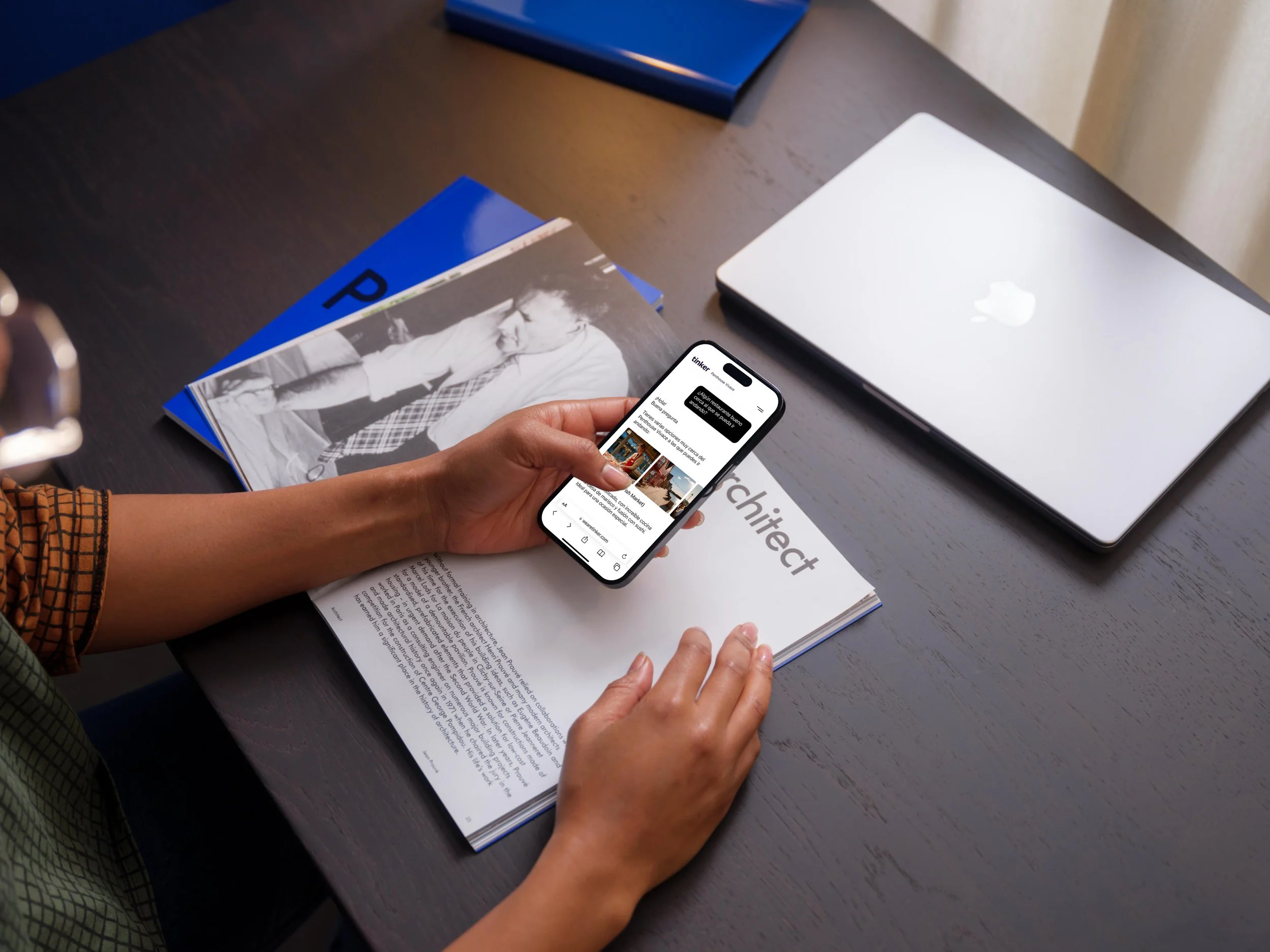 Manos con piel clara sosteniendo un teléfono móvil inteligente mientras revisa contenido en la pantalla, sobre una mesa de color oscuro con una computadora portátil plateada, un libro y un cuaderno azul.