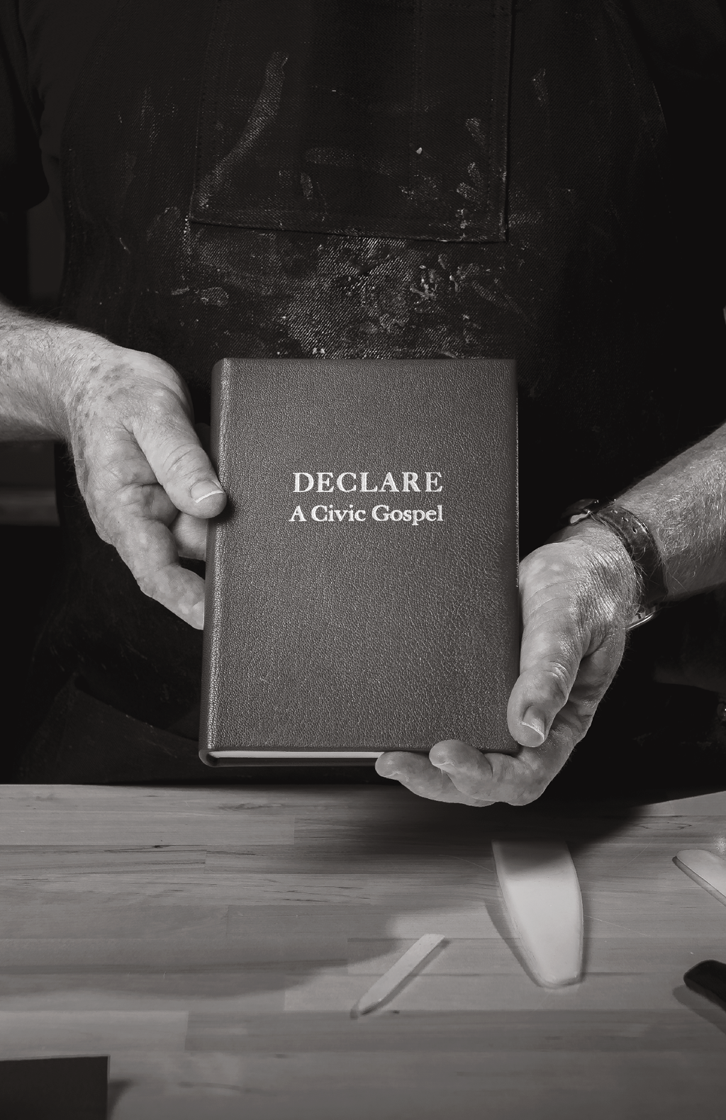 A person holding a Bible titled 'DECLARE: A Civic Gospel' in black and white, with their hands and lower arms visible. The person is wearing a watch on their left wrist and an apron with flour smudges.