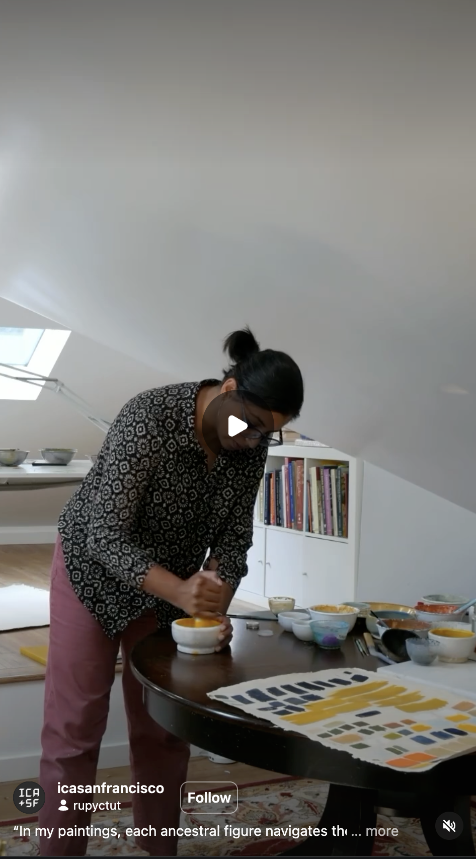 A person with dark hair and glasses is working at a round table with various bowls, paint, and color swatches, in a room with a sloped ceiling, bookshelf, and a skylight window.